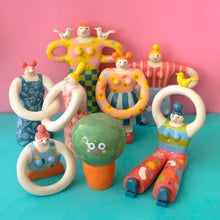 Load image into Gallery viewer, A group of colorful hand-painted ceramic figurines shaped like stylized people with abstract limbs, displayed on a turquoise surface against a pink background.
